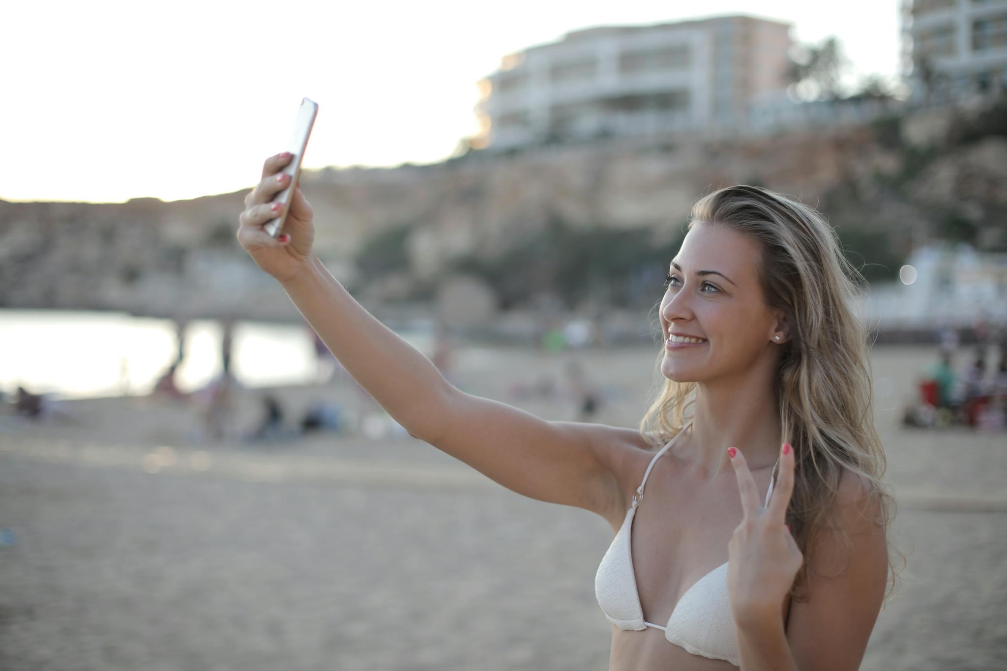 Young woman in bikini posing for a selfie on a sandy beach with a joyful expression.